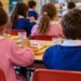 Bambini di spalle con grembiuli rosa e blu pranzano ai tavoli di una mensa scolastica. Immagine generata con intelligenza artificiale