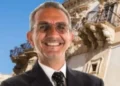 Carmelo Vanasia sorridente in giacca scura e cravatta a pois, con occhiali, davanti a un palazzo barocco con balconi scolpiti