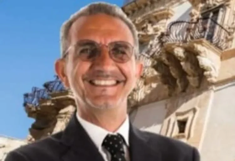 Carmelo Vanasia sorridente in giacca scura e cravatta a pois, con occhiali, davanti a un palazzo barocco con balconi scolpiti