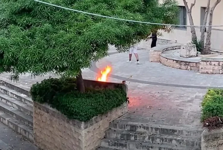 Fiamme su una panchina in pietra accanto a un'aiuola nella piazzetta Sacro Cuore di Comiso, con due persone sullo sfondo