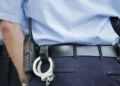 Agente di polizia ripreso di spalle: camicia azzurra, cinturone nero con radio, manette e fondin