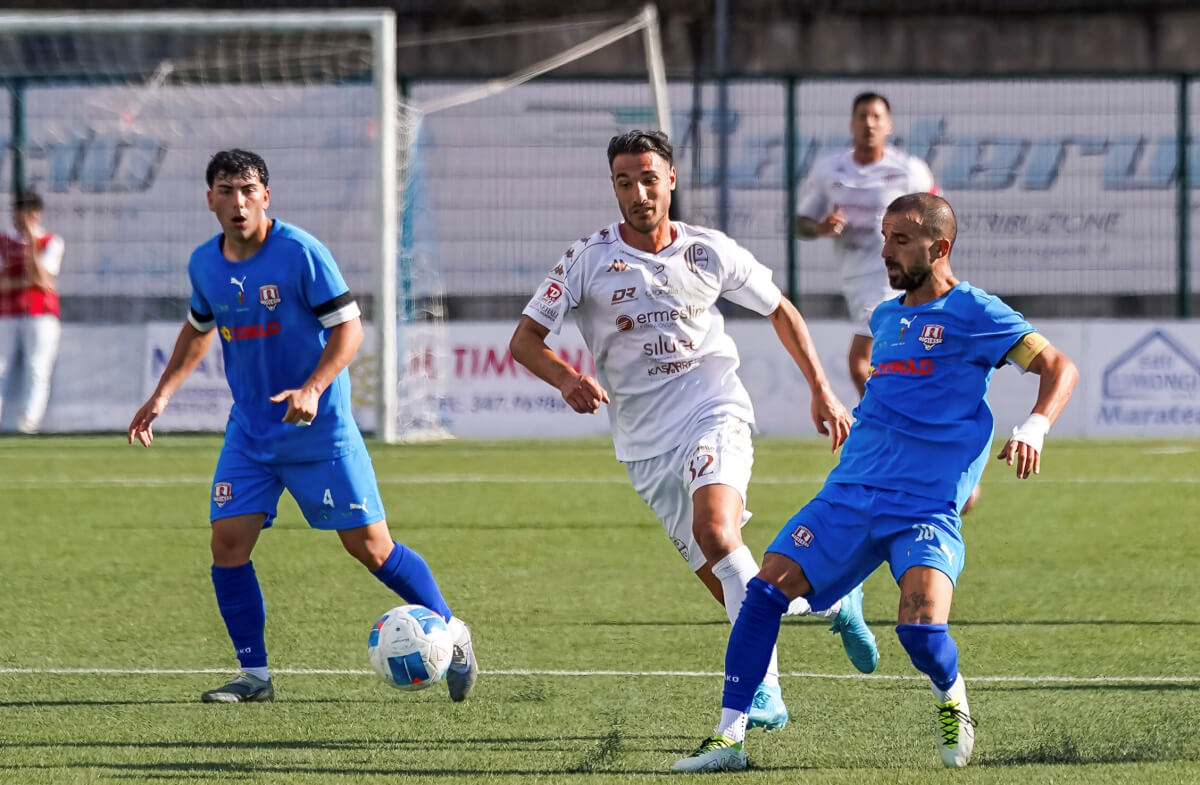 Due calciatori in maglia blu e uno in maglia bianca si contendono il pallone su un campo in sintetico durante Praia-Modica