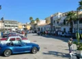 Auto d'epoca parcheggiate in piazza Sorelle Arduino a Scoglitti, tra palme e visitatori, con una cabrio blu in primo piano