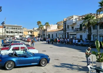 Auto d'epoca parcheggiate in piazza Sorelle Arduino a Scoglitti, tra palme e visitatori, con una cabrio blu in primo piano