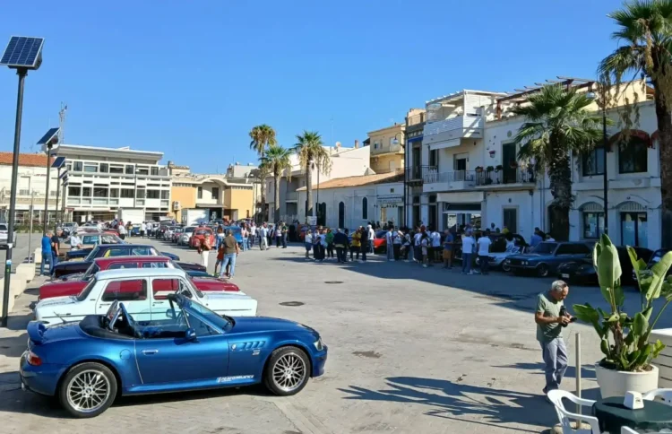 Auto d'epoca parcheggiate in piazza Sorelle Arduino a Scoglitti, tra palme e visitatori, con una cabrio blu in primo piano