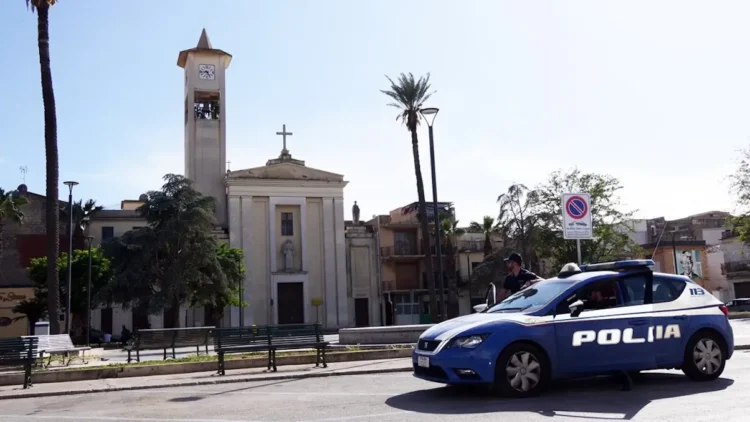 Auto della Polizia di Stato ferma in una piazza di Vittoria, davanti a una chiesa con campanile, palme e panchine