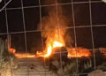 Rifiuti agricoli in fiamme di notte vicino a una serra, con fumo denso dietro una rete metallica a maglie larghe