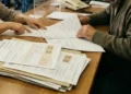Un’impiegata e un contribuente anziano esaminano moduli di tributi e cartelle fiscali su una scrivania allo sportello