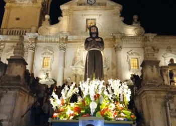 Statua di San Francesco con fiori bianchi e rossi davanti alla facciata barocca illuminata di una chiesa a Ragusa, di sera