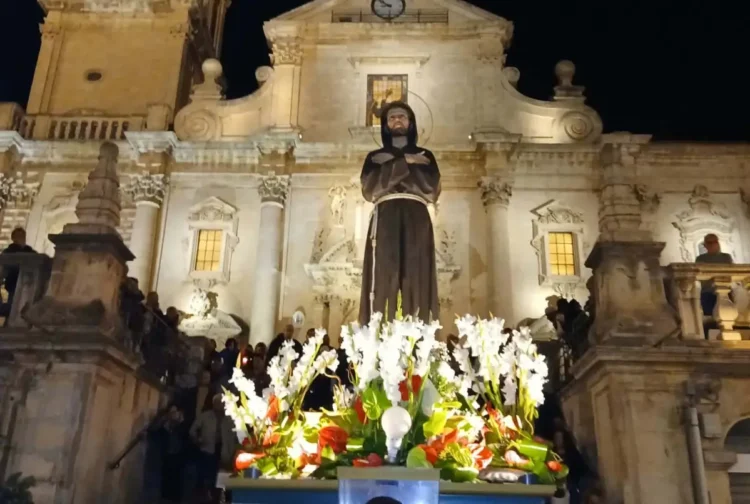 Statua di San Francesco con fiori bianchi e rossi davanti alla facciata barocca illuminata di una chiesa a Ragusa, di sera