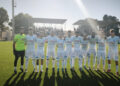 Giocatori del Santa Croce Soccer in maglia bianca schierati in campo con il portiere in verde, tribuna coperta sullo sfondo