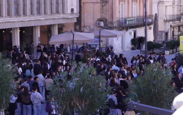 Studenti in sciopero a Ragusa radunati in piazza San Giovanni, tra gli ombrelloni di un bar e piante in primo piano