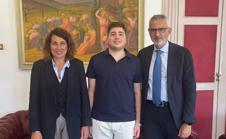 Uno studente con polo blu sorride tra un uomo e una donna in un ufficio, davanti a un dipinto con scene di mietitura