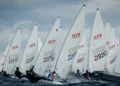 Barche a vela della classe Ilca in regata con mare mosso, velisti sporti fuori bordo e un gommone sulla destra