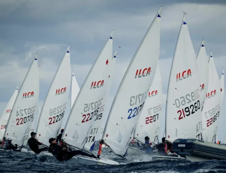 Barche a vela della classe Ilca in regata con mare mosso, velisti sporti fuori bordo e un gommone sulla destra