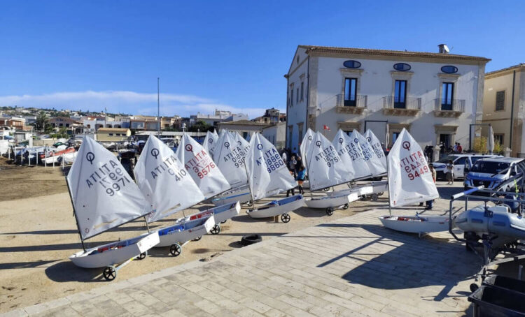 Barche Optimist a terra sul lungomare di Donnalucata durante la Settimana della vela, con le vele alzate davanti a un palazzo