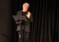 Massimo Leggio legge da un leggio sul palco di Casamatta a Ragusa, davanti a un sipario nero, con una mano alzata