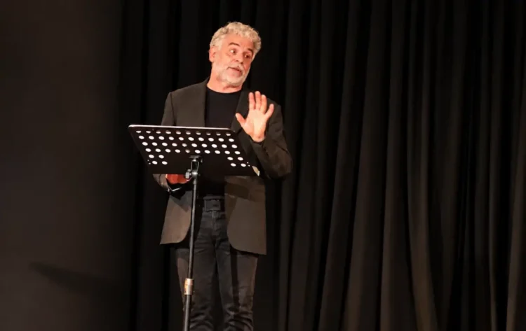 Massimo Leggio legge da un leggio sul palco di Casamatta a Ragusa, davanti a un sipario nero, con una mano alzata