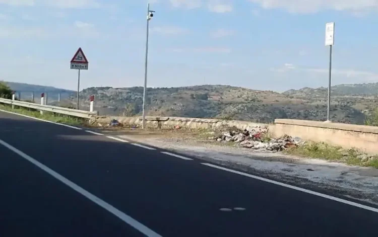 Telecamera su palo in una piazzola della Ss 194 a Ragusa, con rifiuti abbandonati vicino al muretto e colline sullo sfondo