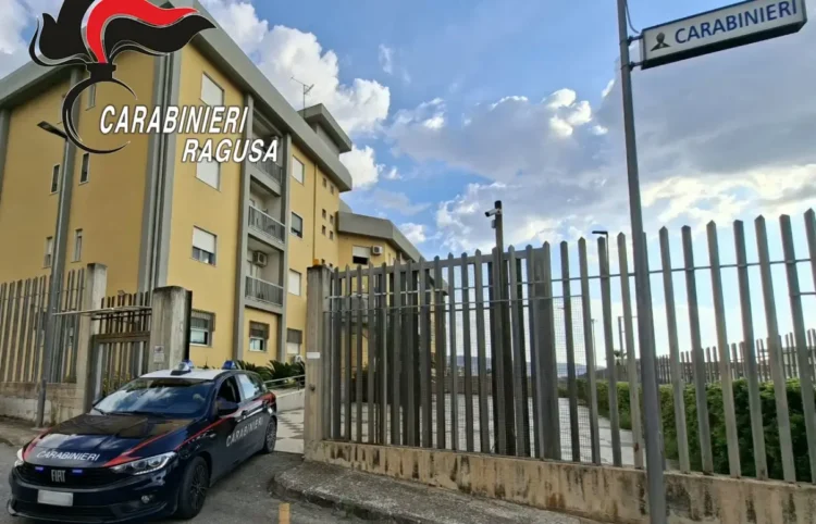 Auto dei carabinieri parcheggiata davanti al cancello di una caserma, con un palazzo giallo e il cartello «Carabinieri»