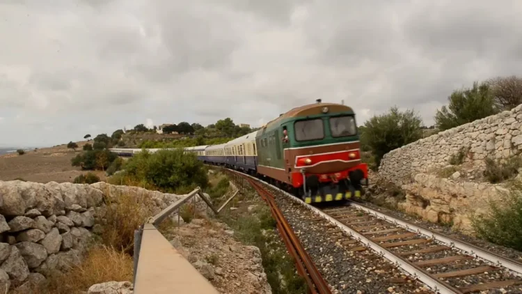 Treno storico con locomotiva diesel e carrozze d'epoca blu e crema in curva tra muretti a secco e macchia mediterranea
