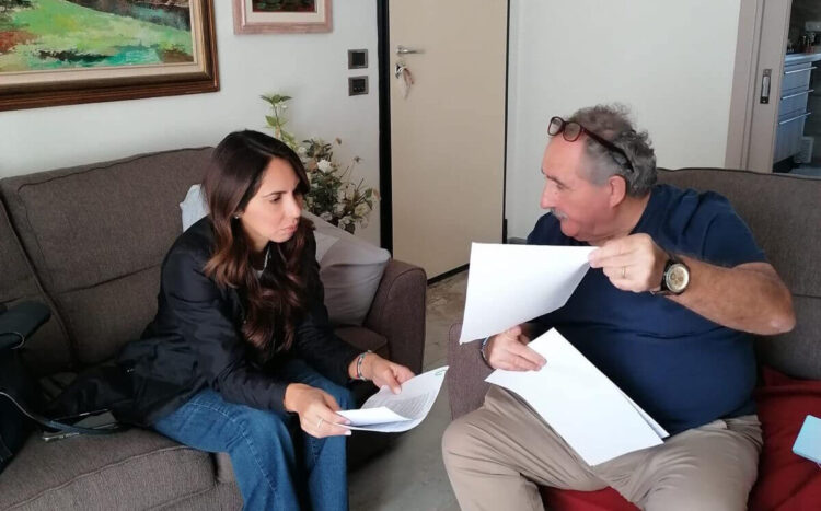 Rossana Caruso legge un foglio seduta su un divano accanto a un uomo con occhiali sulla testa che le mostra dei documenti