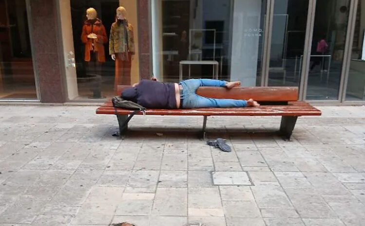 Persona distesa a dormire su una panchina di legno in via Roma a Ragusa, davanti alla vetrina di un negozio di abbigliamento