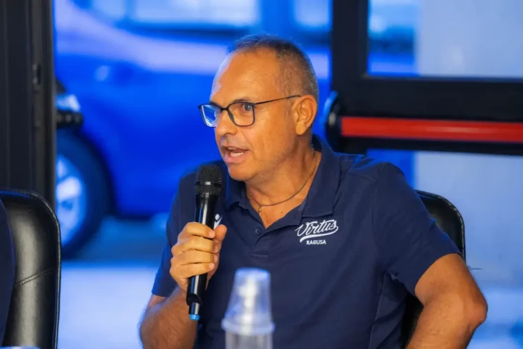 Il coach della Virtus Ragusa Gianni Recupido parla al microfono con polo blu e occhiali, alla vigilia della trasferta a Catanzaro